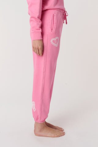 A person wears the Roller Rabbit Vintage Valentine Sweatpants—pink fleece with a drawstring waist and white heart designs—standing barefoot against a plain light background, showcasing only the lower half of their body.
