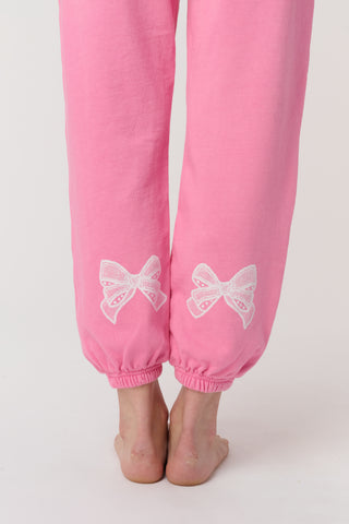 A person wears the Roller Rabbit Vintage Valentine Sweatpants—pink fleece with a drawstring waist and white heart designs—standing barefoot against a plain light background, showcasing only the lower half of their body.