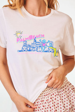 A smiling woman wears a Roller Rabbit Greetings From Palm Beach Tee, an unlined, cotton jersey pull-over t-shirt featuring a colorful graphic of a Palm Beach skyline with a sun and a palm tree.