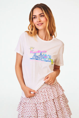 A smiling woman wears a Roller Rabbit Greetings From Palm Beach Tee, an unlined, cotton jersey pull-over t-shirt featuring a colorful graphic of a Palm Beach skyline with a sun and a palm tree.