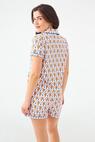 A woman with long brown hair models the Roller Rabbit Campus Lovely Monkey Polo Pajamas—an orange and navy animal print short-sleeved button-up top with matching shorts—against a plain light backdrop.