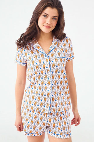 A woman with long brown hair models the Roller Rabbit Campus Lovely Monkey Polo Pajamas—an orange and navy animal print short-sleeved button-up top with matching shorts—against a plain light backdrop.
