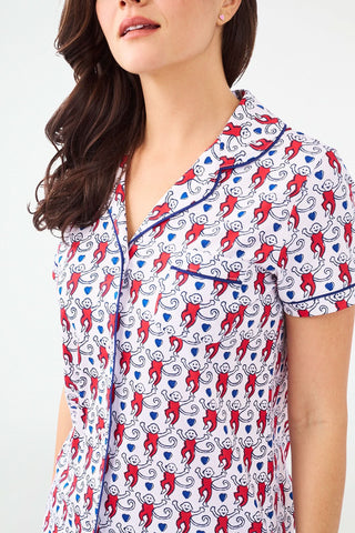 A woman stands with hands behind her back in Roller Rabbit's Campus Lovely Monkey Polo Pajamas, a short-sleeved, button-up red and navy romper with playful patterns—perfect sleepwear for showing your school spirit.