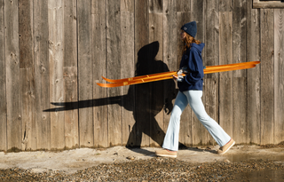 Person carrying orange skis against a wooden wall