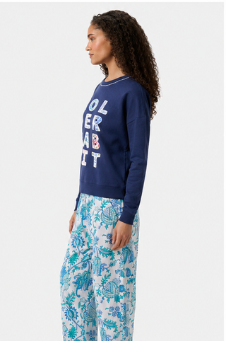 A woman with curly hair stands in profile against a plain background, wearing the Roller Rabbit Applique Crewneck by Roller Rabbit and loose white pants with blue and green floral patterns.